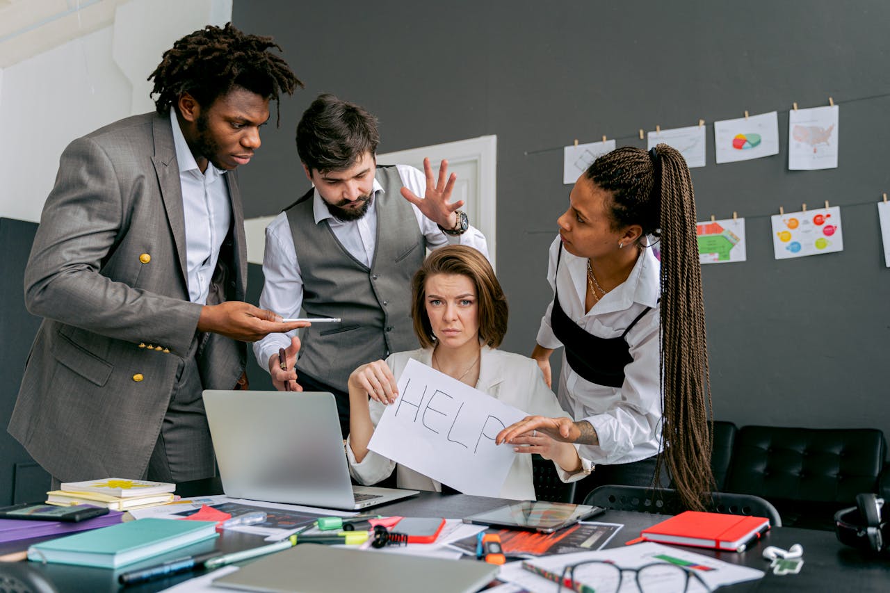home-services-03 Office scene with diverse colleagues around a stressed woman holding a sign that says HELP. Focus on teamwork dynamics.